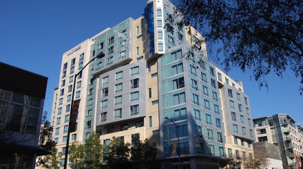 Modern hotel with a glass facade and colorful accents, surrounded by trees and city buildings under a clear blue sky.